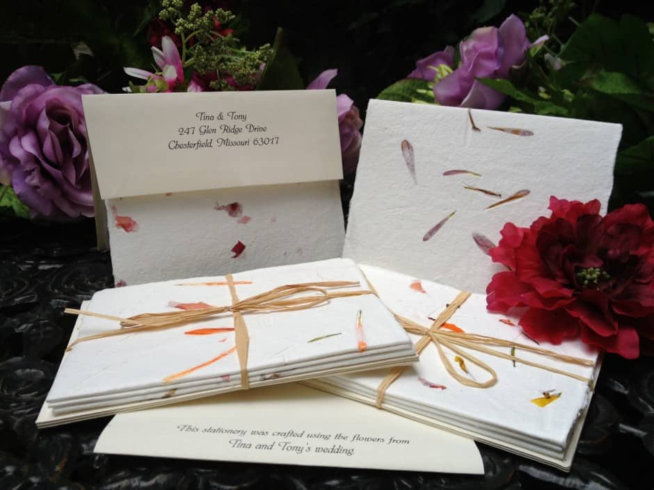 Handmade wedding invitations and envelopes with natural flower petal inclusions, displayed with fresh flowers on a dark surface. a placard provides details about the craftsmanship and origin.