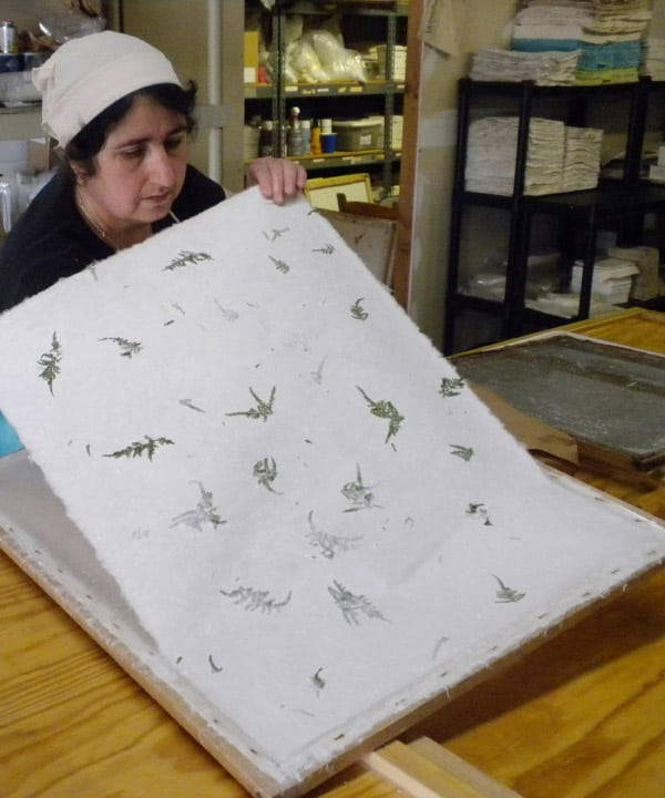 A woman in a workshop holding up a large sheet of handcrafted papers with green botanical prints. She is examining the paper's texture and quality.