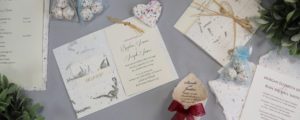 A flat lay of wedding invitations made from seed paper, surrounded by decorative elements like eucalyptus leaves, twine, and patterned hearts on a soft gray background.