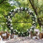 An outdoor wedding setup with a large floral arch of white and green flowers, surrounded by wooden logs, white chairs, and scattered petals, set against a lush green backdrop.