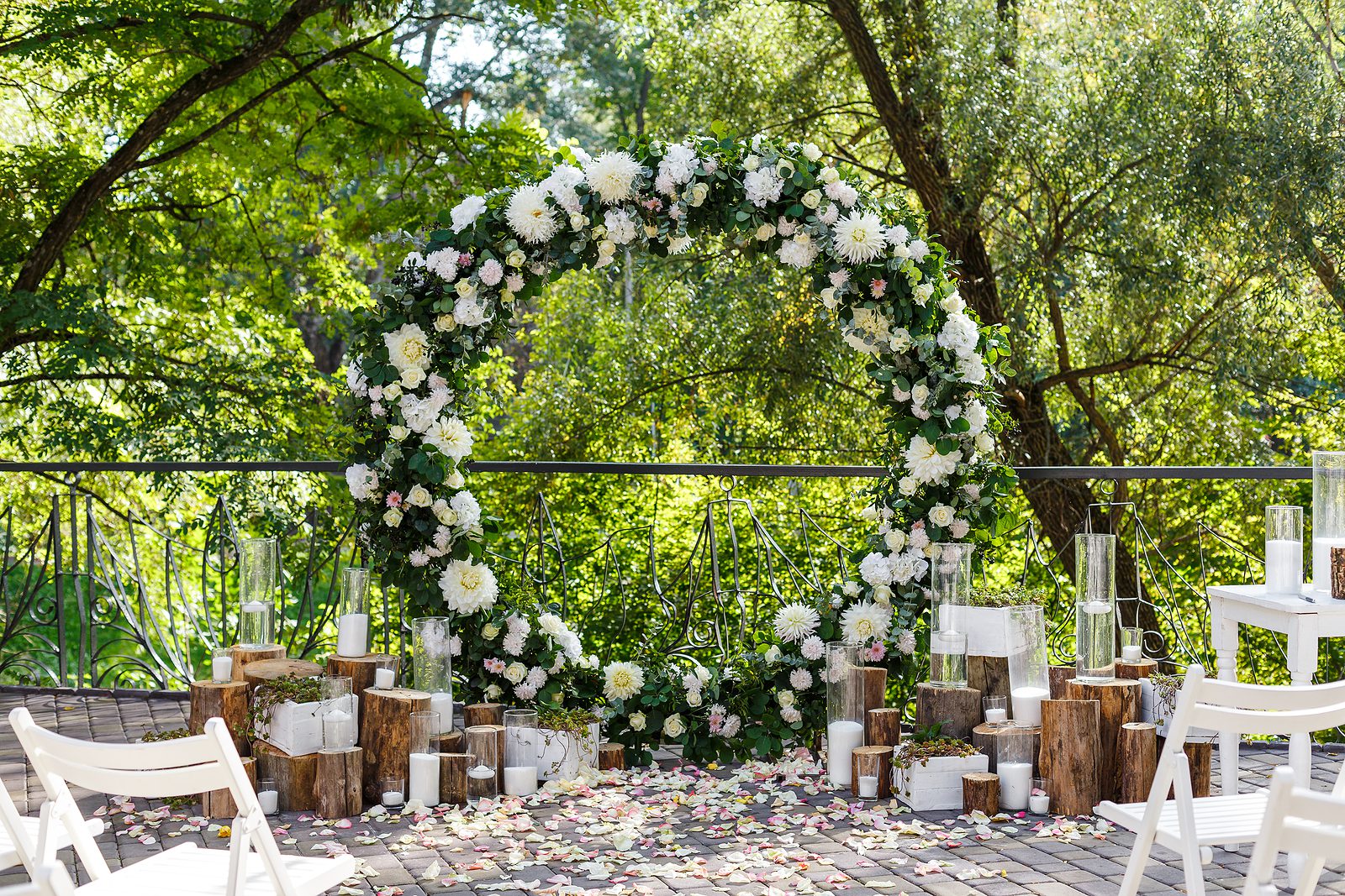 An outdoor wedding setup with a large floral arch of white and green flowers, surrounded by wooden logs, white chairs, and scattered petals, set against a lush green backdrop.