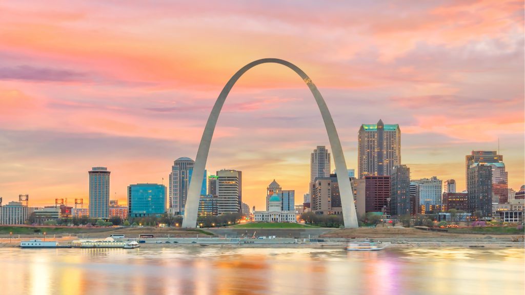 A scenic view of the st. louis skyline at sunset, featuring the gateway arch and color-drenched clouds reflected off the mississippi river, with boats floating on the water.