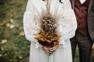 A bride in a lace dress and a groom in a tweed suit hold a bouquet of autumn leaves and pampas grass, focusing on their midsections with a green grassy background.