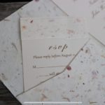 Close-up of a wedding rsvp card crafted from handmade paper with visible flower petals, requesting a reply by august 1st.
