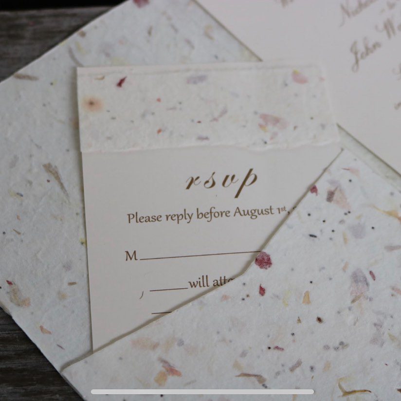 Close-up of a wedding rsvp card crafted from handmade paper with visible flower petals, requesting a reply by august 1st.