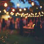 Outdoor evening party with string lights hanging between trees, creating a warm, festive ambiance. Blurred figures of people are mingling in the background, while greenery is visible in the foreground.