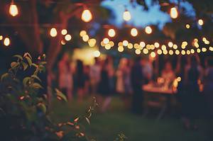 Outdoor evening party with string lights hanging between trees, creating a warm, festive ambiance. Blurred figures of people are mingling in the background, while greenery is visible in the foreground.