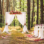 An outdoor wedding setup in a forest with tall trees. A white fabric arch adorned with pink and orange flowers stands at the aisle's end. White chairs draped with fabric and decorated with flowers line both sides, with pink flower petals scattered along the pathway.