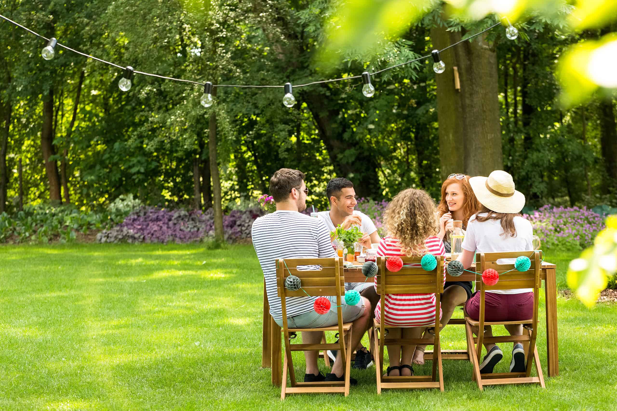 A group of five people sit around a wooden table outdoors, enjoying drinks in a grassy area. The table is adorned with colorful decorations, and there are string lights overhead. The setting is lush with greenery, and it appears to be a sunny day.