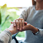A close-up of two people holding hands in a comforting manner. One person is gently grasping the other's hand with both of theirs. The background is blurred with greenery, suggesting an indoor setting with plants.