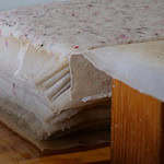 A stack of handmade paper sheets with visible fibers and flower petals, resting on a wooden surface, likely during the papermaking process.