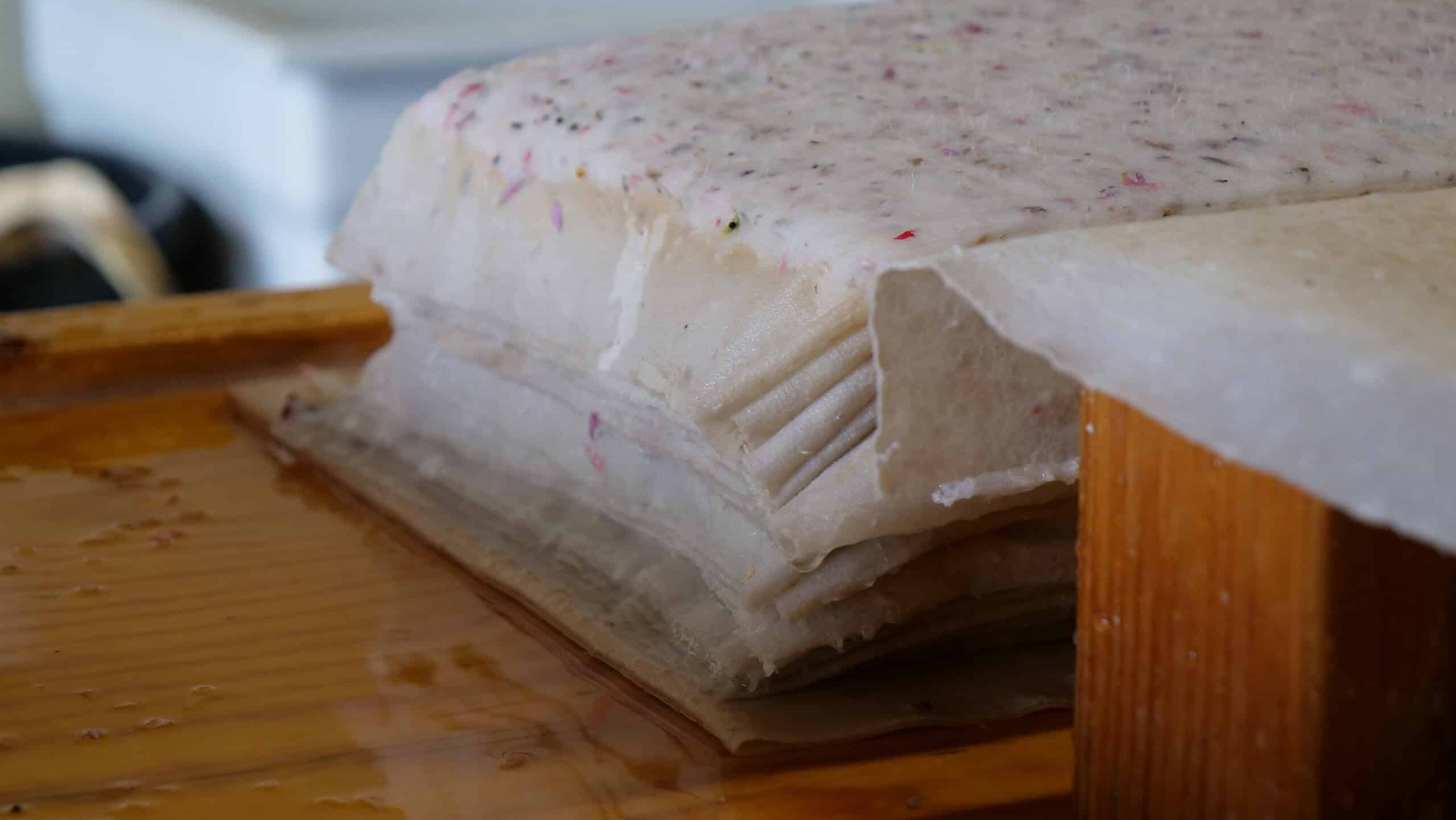 A stack of handmade paper sheets with visible fibers and flower petals, resting on a wooden surface, likely during the papermaking process.