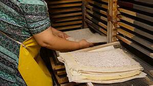 A person wearing a patterned shirt and yellow apron places handmade paper sheets on racks to dry, with stacks of paper and wooden drying trays visible.